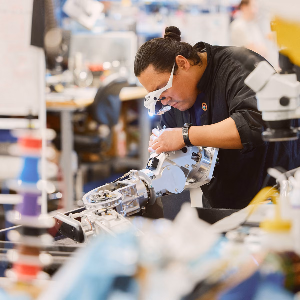 Technician wearing magnifying glasses with a light closely inspects and works on a metallic robotic arm in a workshop filled with tools and equipment.