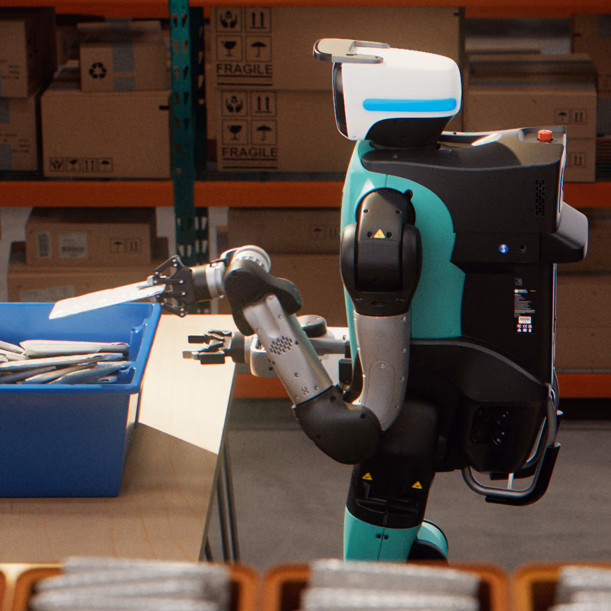 A teal and black humanoid robot with a white head is handling items in a blue bin while positioned next to a table in a warehouse environment with shelves and cardboard boxes labeled "fragile" in the background.