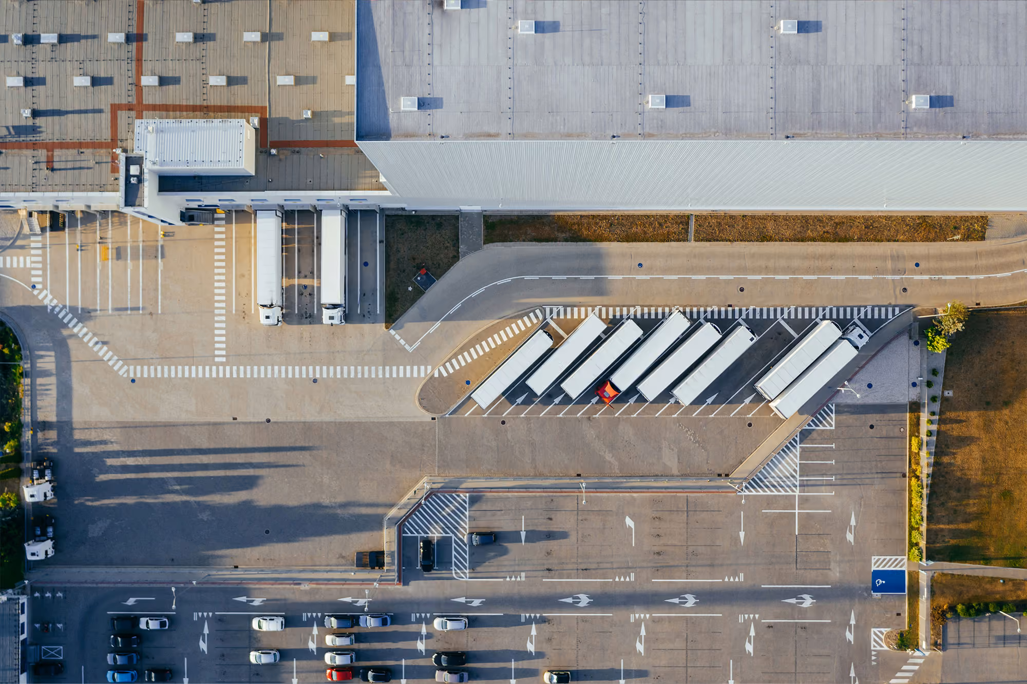 Aerial view of an industrial facility with a large building, several parked semi-truck trailers in designated bays, and multiple cars in a parking lot with marked lanes and crosswalks.
