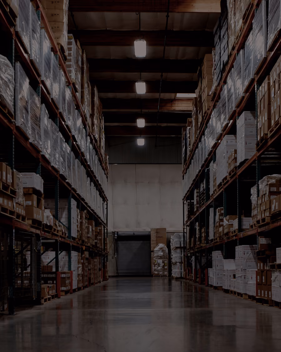 Interior of a large warehouse with tall metal shelves filled with stacked boxes and pallets lining both sides of a wide aisle, illuminated by ceiling lights.