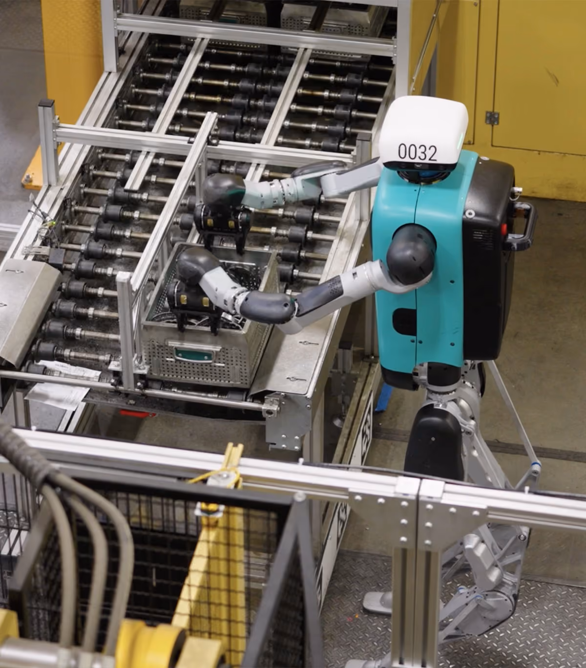 A teal and silver robot with the number 0032 uses its two articulated arms to handle a gray metal basket on a conveyor system in a factory setting.