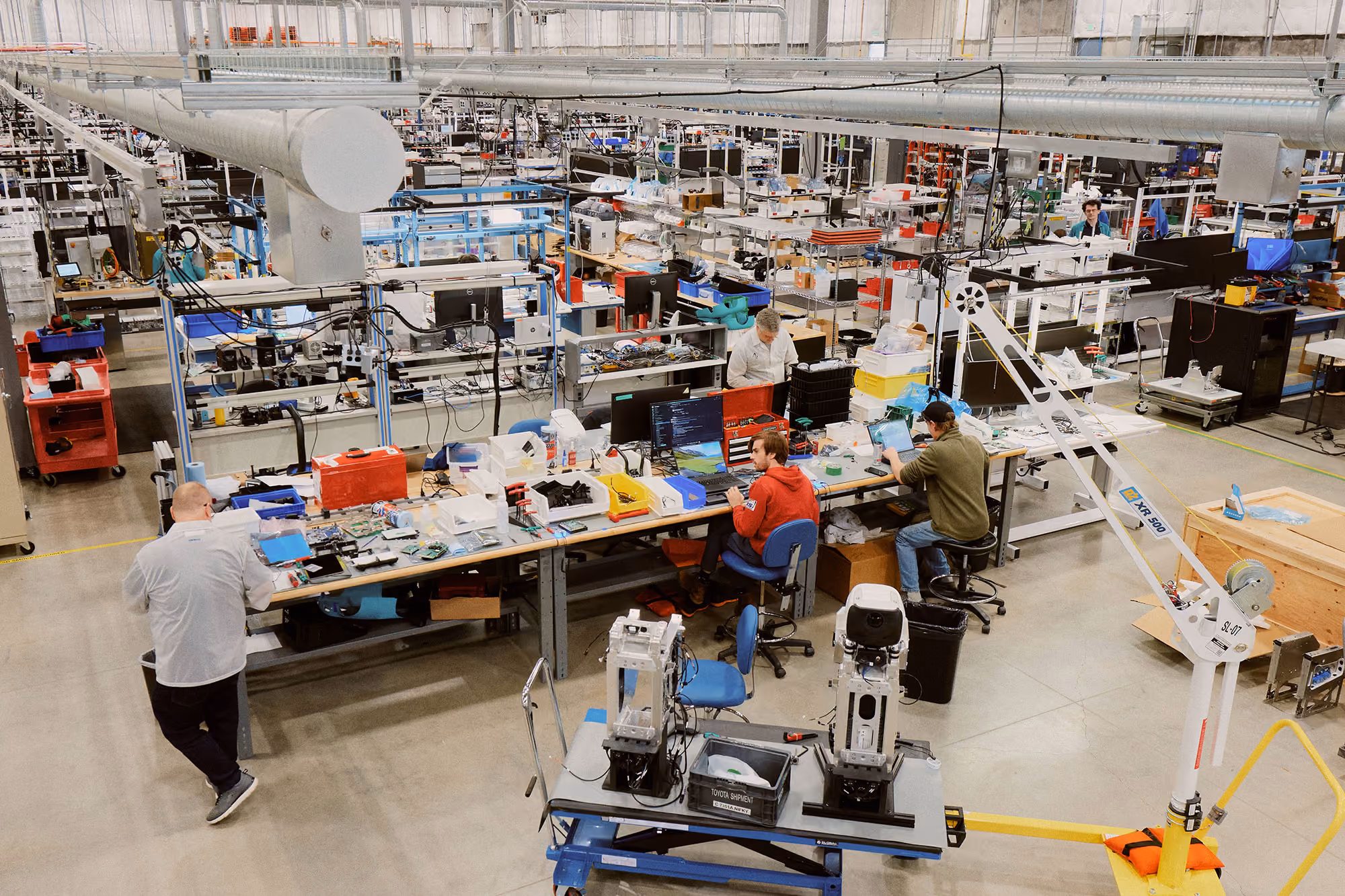 Wide view of a spacious assembly workshop with workers seated and standing at tables filled with components and tools, and robotic devices on a table in the foreground.