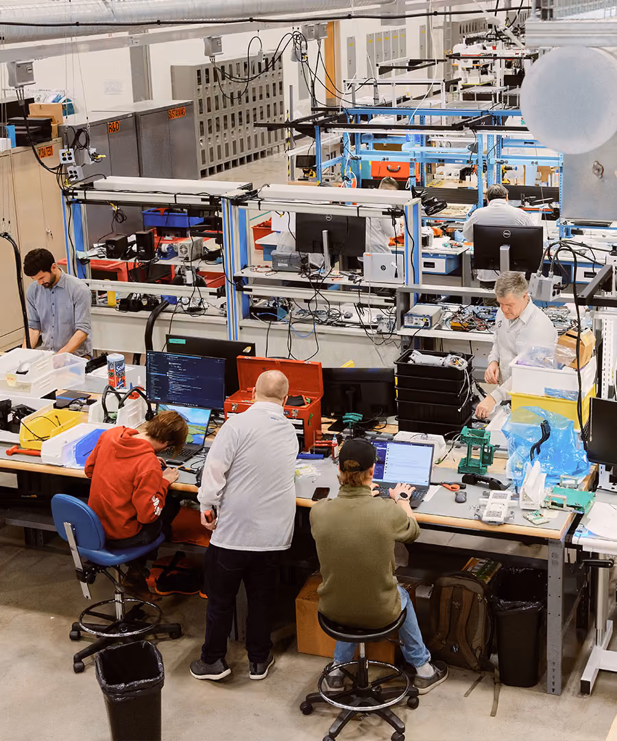 A busy electronics workshop with six people working at long tables filled with multiple computer screens, laptops, electronic devices, tools, and wiring in a large industrial space.