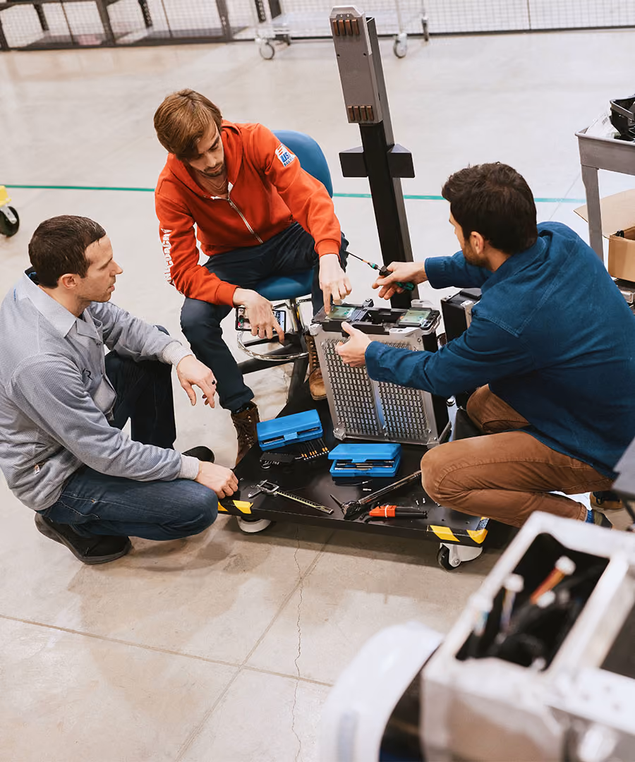 Three men working collaboratively on a piece of electronic equipment indoors, surrounded by tools and parts on a wheeled platform.