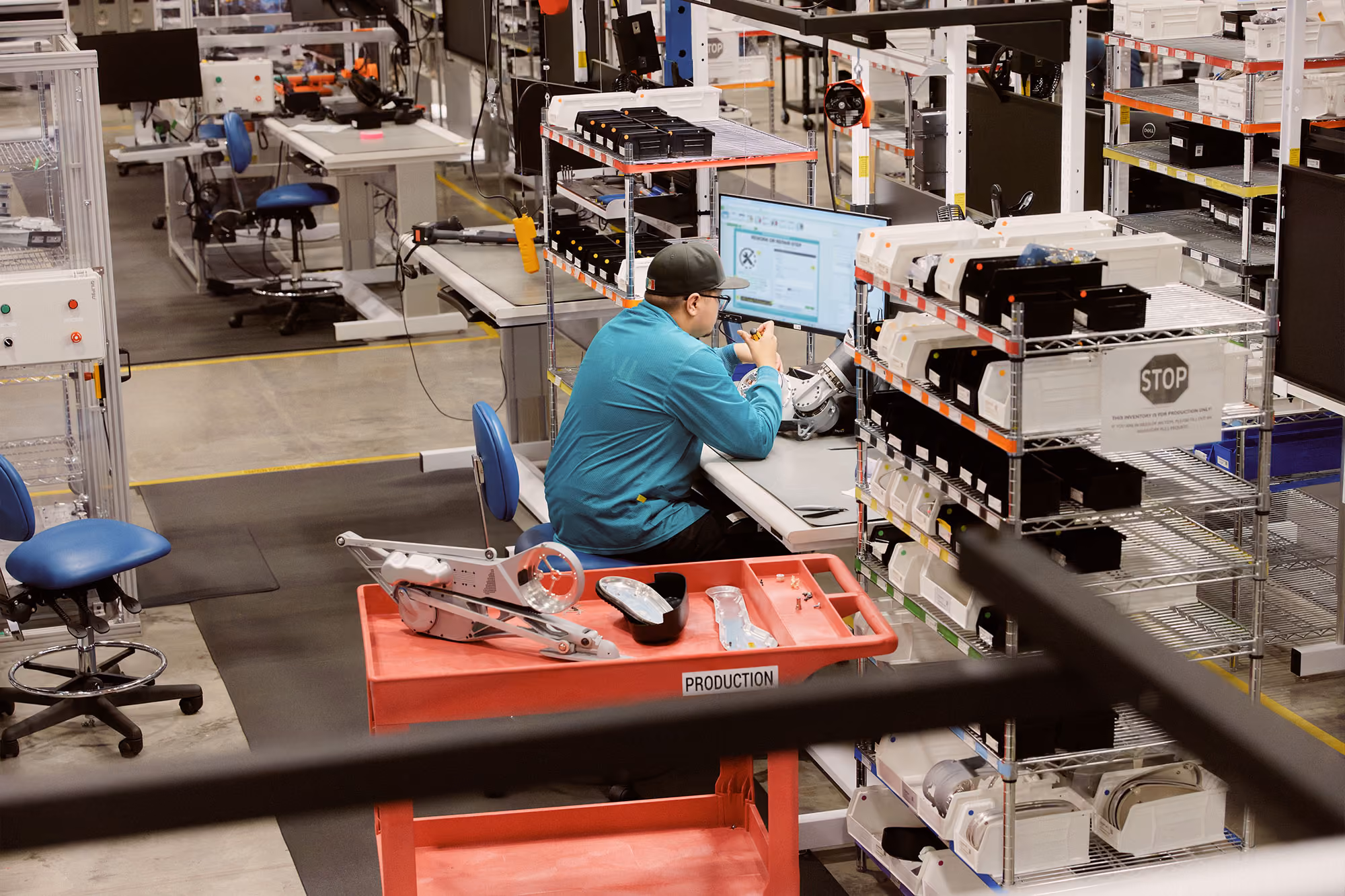 A worker wearing a teal long-sleeve shirt, black cap, and glasses is seated at a workstation in a production facility, examining a small part under a bright light, surrounded by shelves with organized components and a red cart labeled 'PRODUCTION' with various parts.