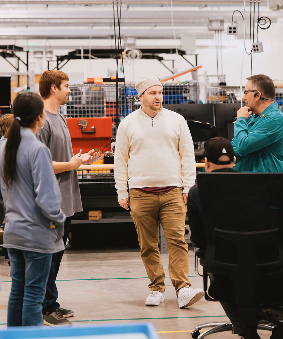Five people engaged in a discussion inside an industrial or workshop setting with machinery and equipment in the background; one person in a white sweater and knit hat is central and speaking while others listen.