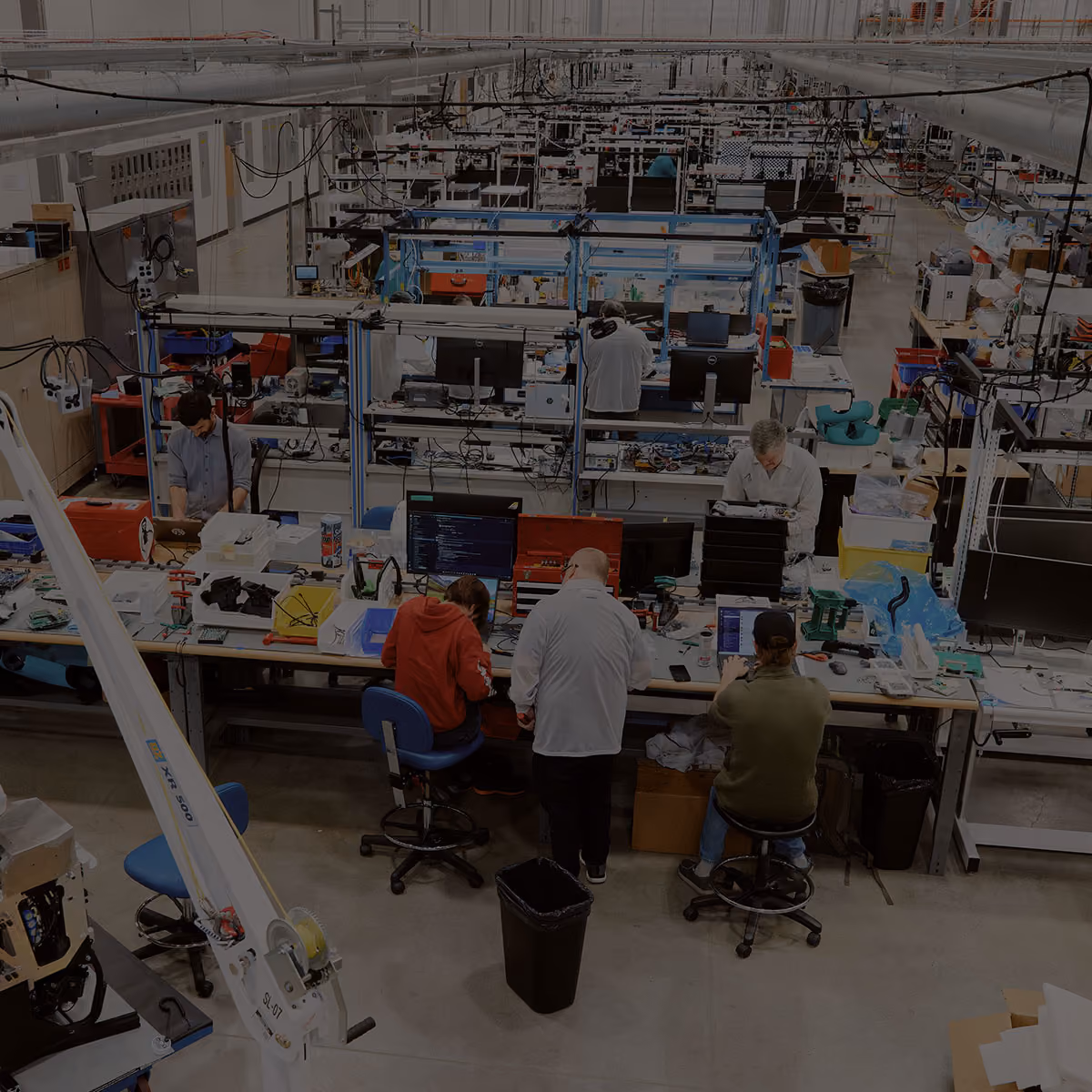 A spacious tech workshop with multiple workstations where several people are working on computers and electronics, surrounded by tools and equipment.