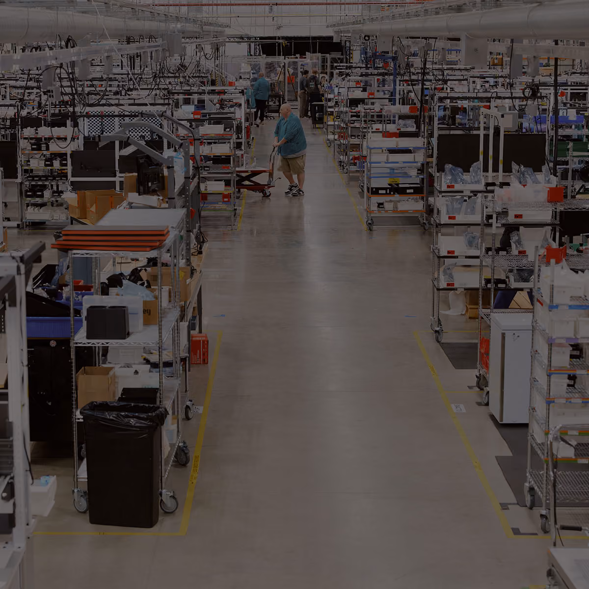 Interior of a large manufacturing facility with multiple rows of metal shelving units and workstations, workers in casual attire moving and working among the shelves, with a wide central aisle and overhead lighting.