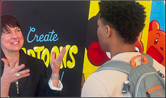 A woman gestures while speaking to a young man with a backpack in front of a colorful wall with the words 'Create Cartoons'.