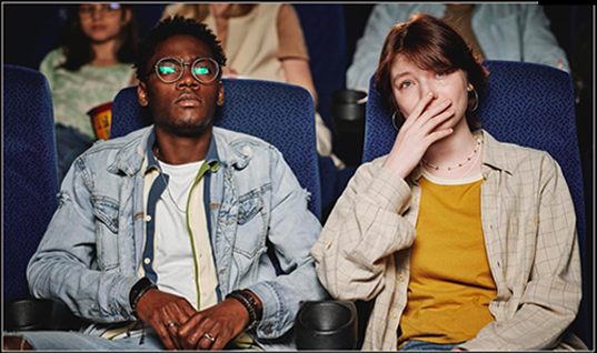 Two people seated in a dark movie theater, one wearing glasses and a denim jacket, the other covering their mouth with a hand.