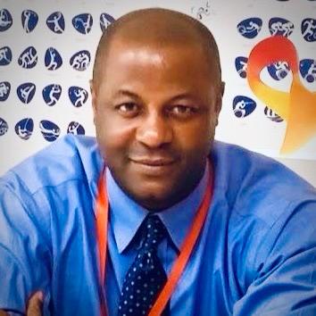 Man in blue shirt and dark tie sitting in front of a backdrop with blue and white logos.