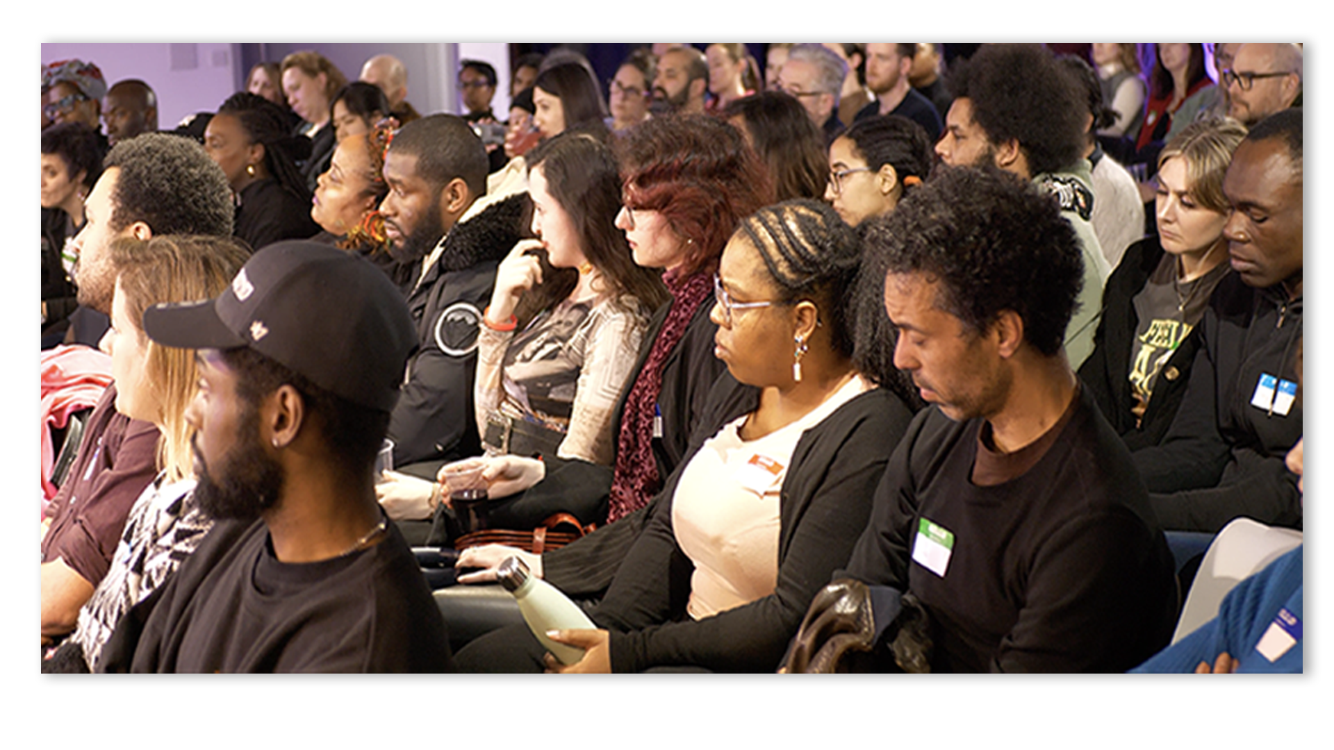 A diverse group of adults seated closely in rows, attentively listening at an indoor event.
