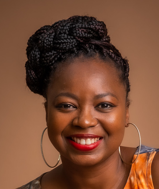 Smiling woman with braided hair styled in an updo, wearing large hoop earrings and red lipstick against a plain brown background.