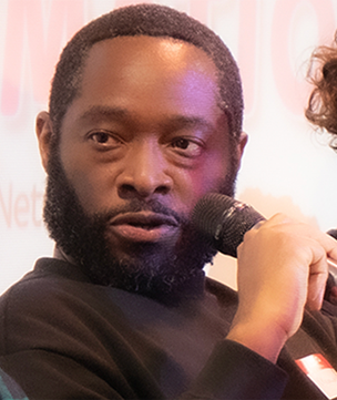 Man with a beard holding a microphone speaking, with partial view of another person on the right.