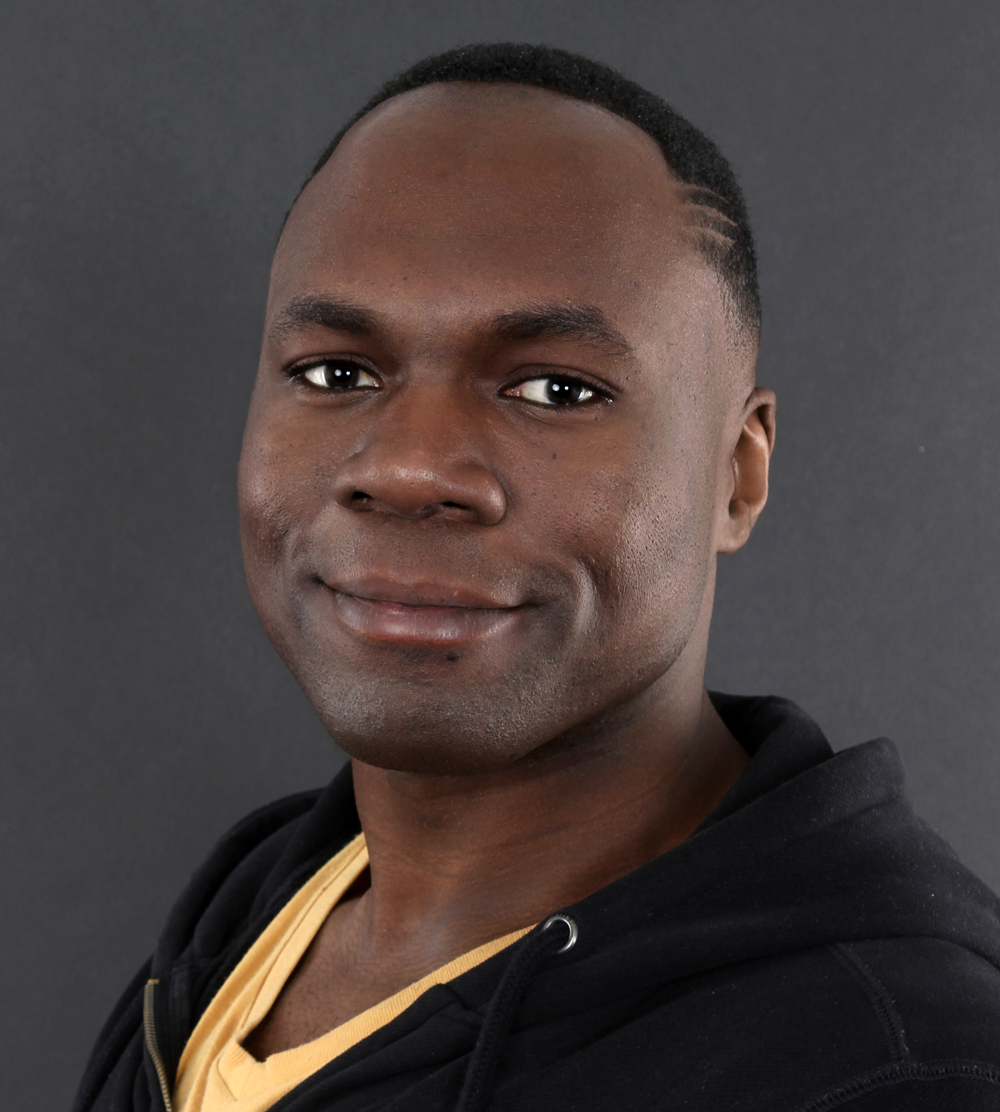 Smiling man with short hair wearing a yellow shirt and black hoodie against a dark gray background.