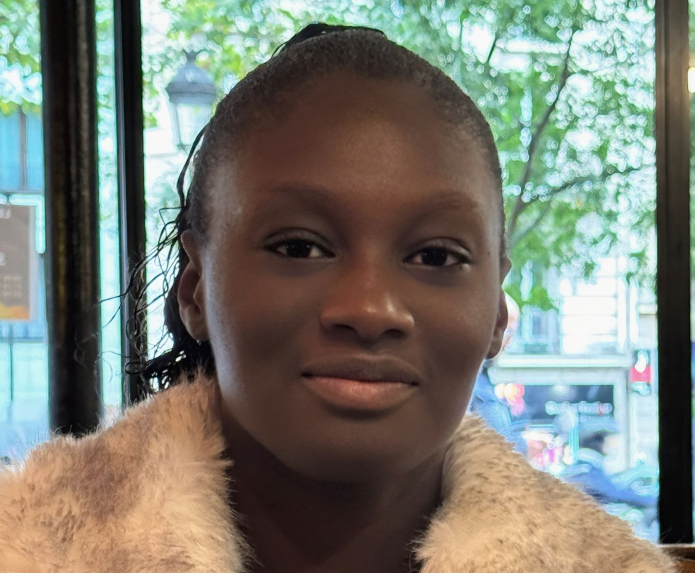 Close-up portrait of a woman with dark skin wearing a light-colored fur coat, with a blurred window and green trees in the background.