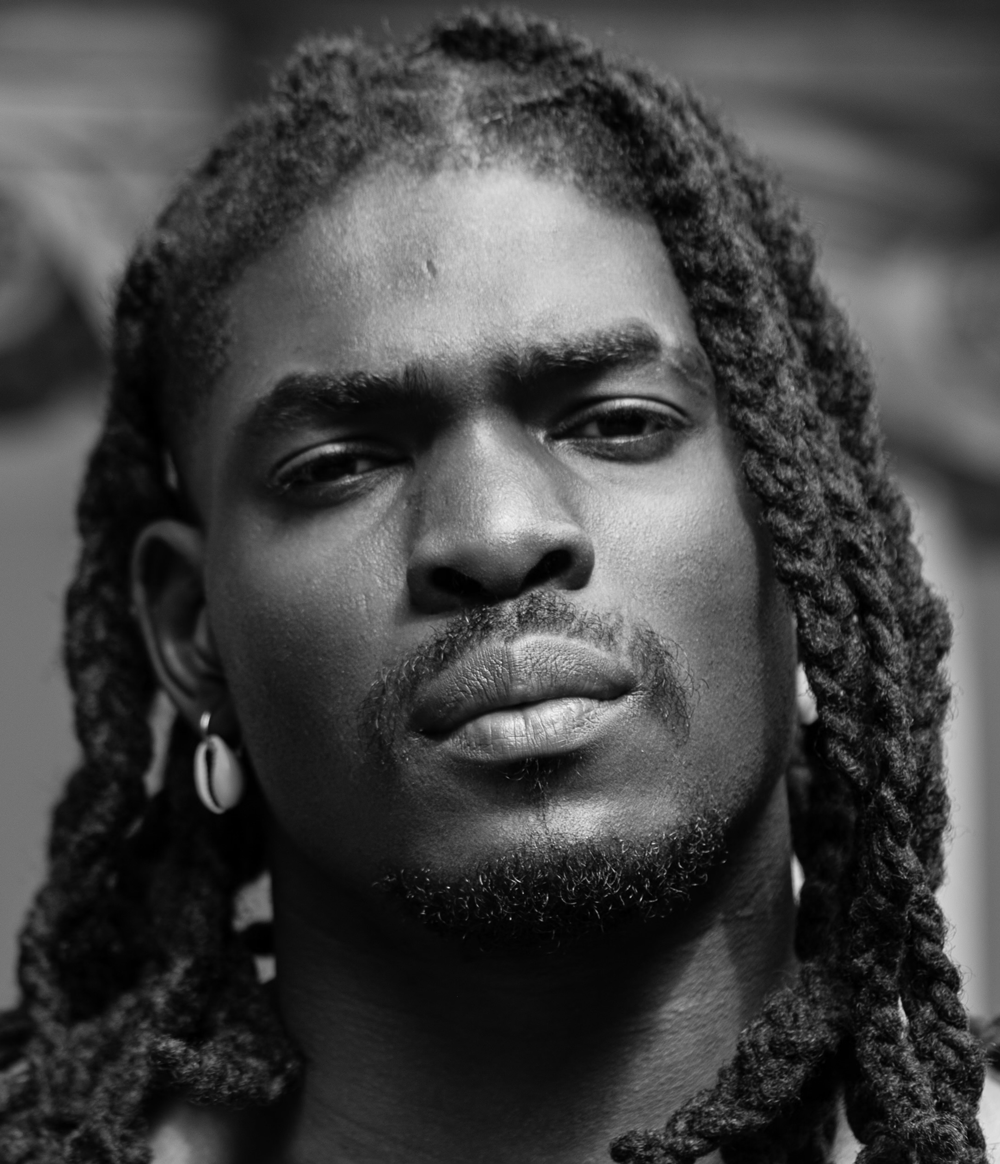 Close-up black and white portrait of a man with dreadlocks and hoop earrings looking directly at the camera.