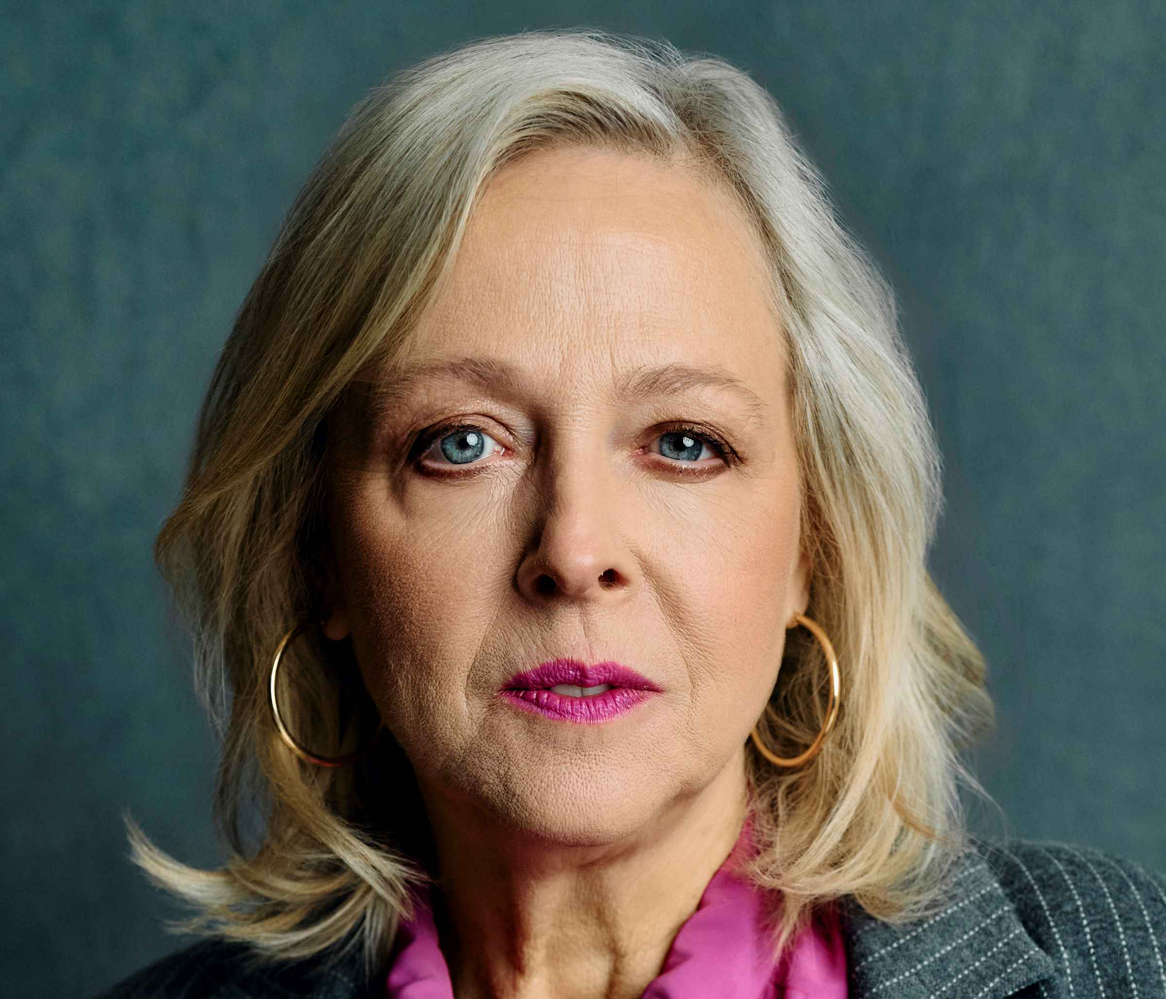 Close-up portrait of a middle-aged woman with gray hair, blue eyes, pink lipstick, gold hoop earrings, and wearing a gray pinstripe jacket with a pink ruffled blouse.