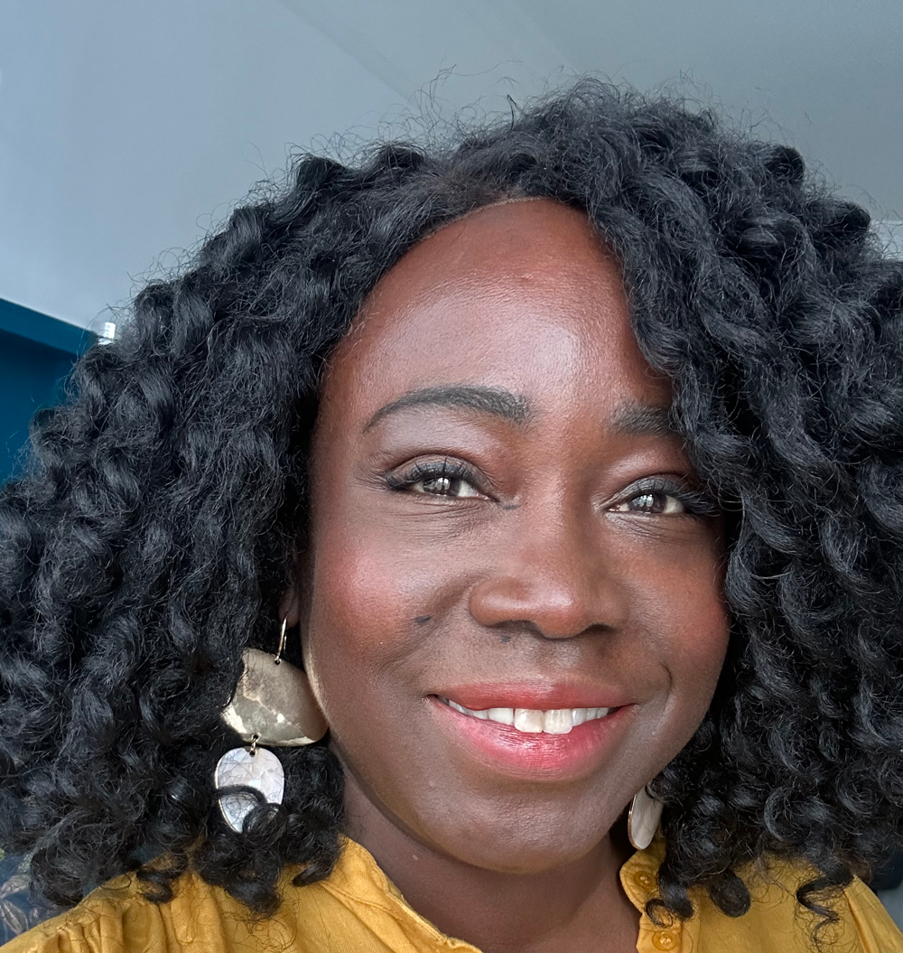 Smiling woman with curly black hair wearing large metallic earrings and a mustard yellow top.