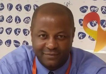 Man in a blue shirt with a red lanyard, smiling at the camera against a backdrop featuring various sports icons and a ribbon symbol.