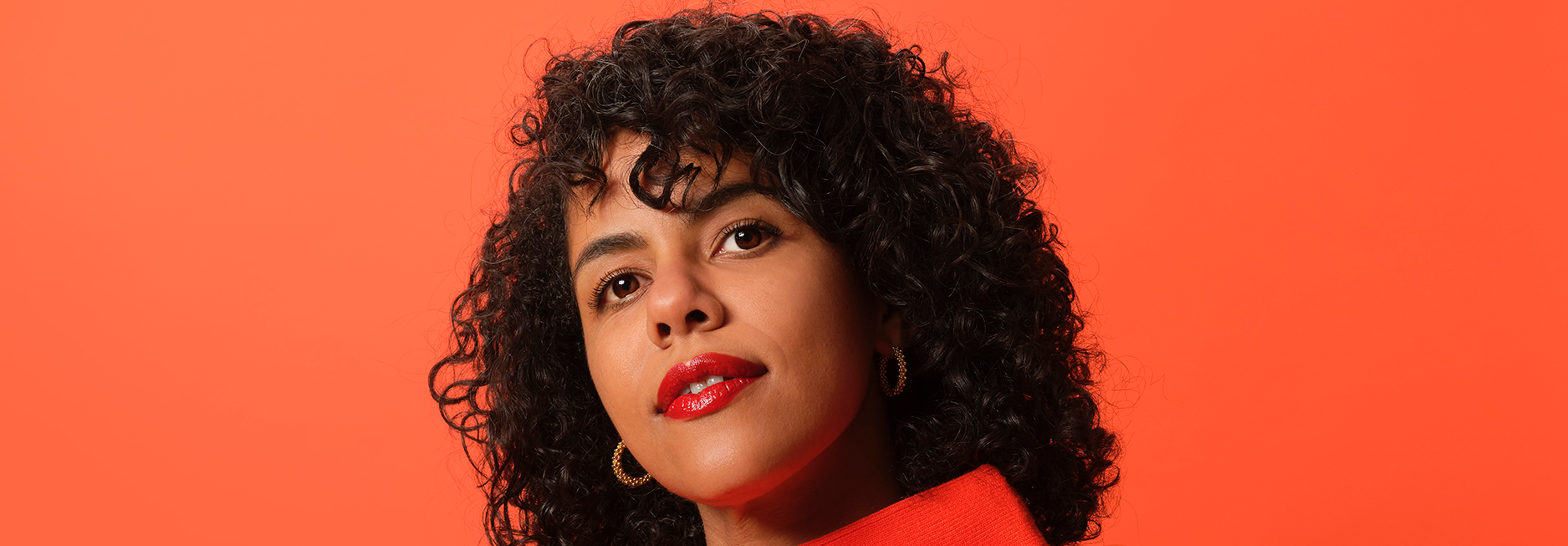 Woman with curly black hair wearing red lipstick, gold hoop earrings, and a red top against an orange background.