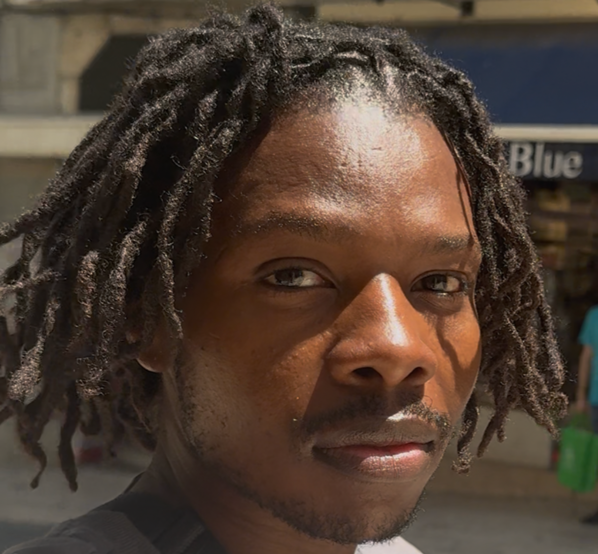 Close-up of a man with medium-length dreadlocks looking at the camera in natural sunlight.