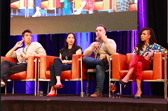 Four panelists sitting on orange chairs on a stage during a discussion, one man speaking into a microphone.