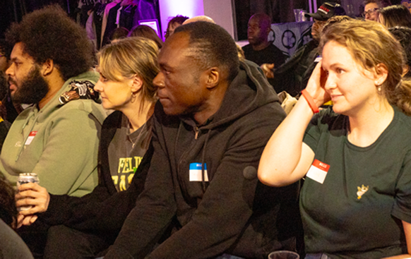 A diverse group of four people sitting closely together, attentively watching something off-camera in an indoor setting.