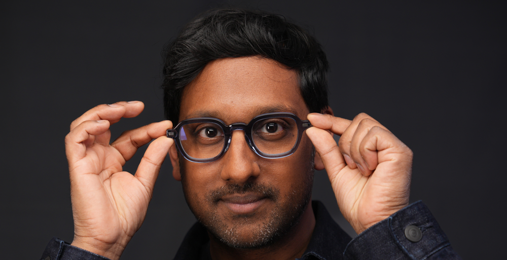 Man with black hair adjusting his dark-framed glasses against a dark background.
