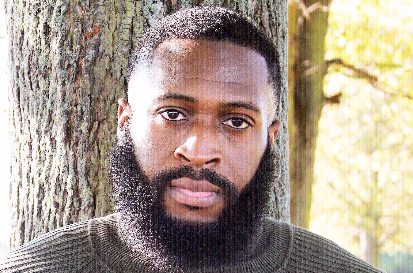 Portrait of a man with a full beard and short hair leaning against a tree trunk outdoors.