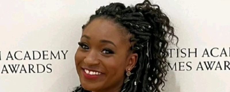 Smiling woman with braided hair styled in a high ponytail, standing in front of a British Academy Games Awards backdrop.