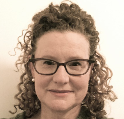 Close-up of a smiling woman with curly hair, wearing glasses and pearl earrings, against a plain light background.