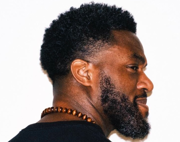 Side profile of a man with a full beard, short curly hair, and wearing a wooden bead necklace and black shirt.