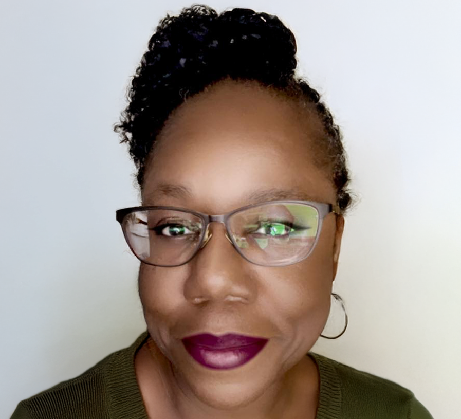 Close-up of a smiling woman wearing glasses, dark purple lipstick, hoop earrings, and a green sweater against a plain background.