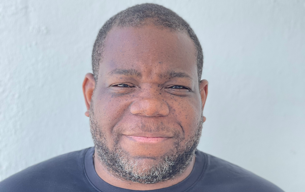 Close-up portrait of a smiling man with short hair and a salt-and-pepper beard wearing a dark shirt against a light gray background.