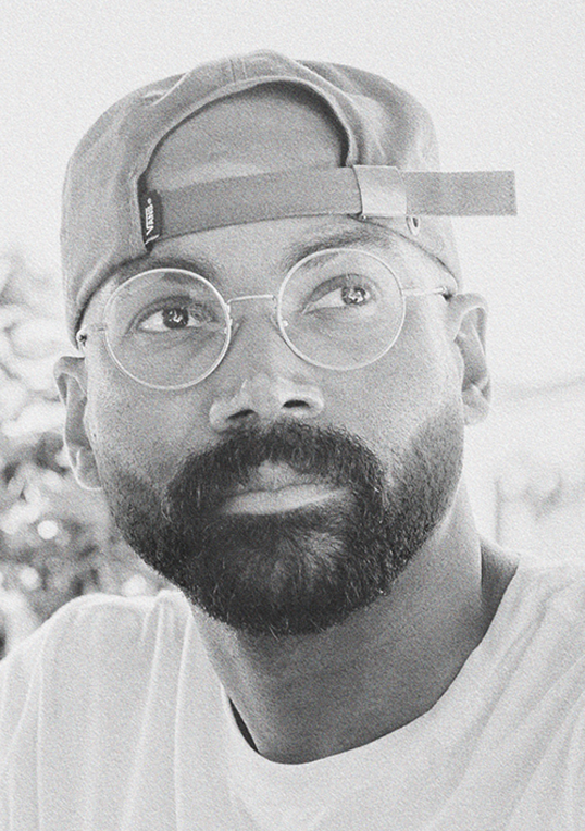 Black man with a beard wearing round glasses and a backward baseball cap looking thoughtfully to the side.