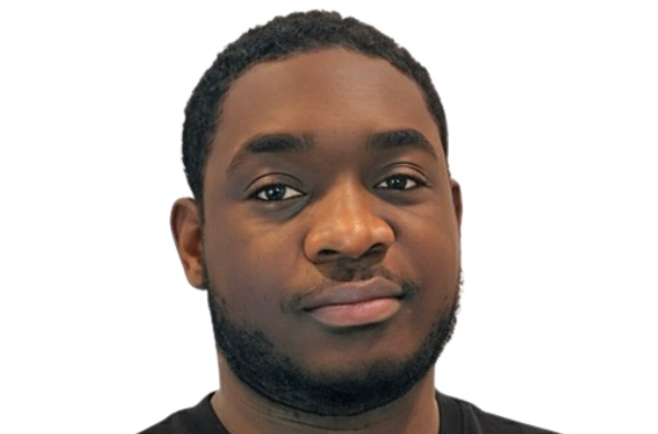 Portrait of a young man with short curly hair and a beard wearing a black shirt against a white background.