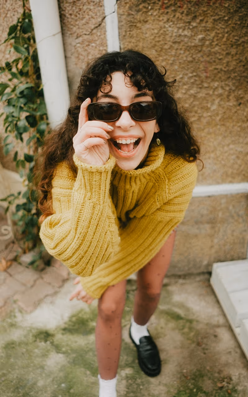 Happy young woman with curly hair wearing a yellow sweater and sunglasses, smiling and leaning forward outdoors.