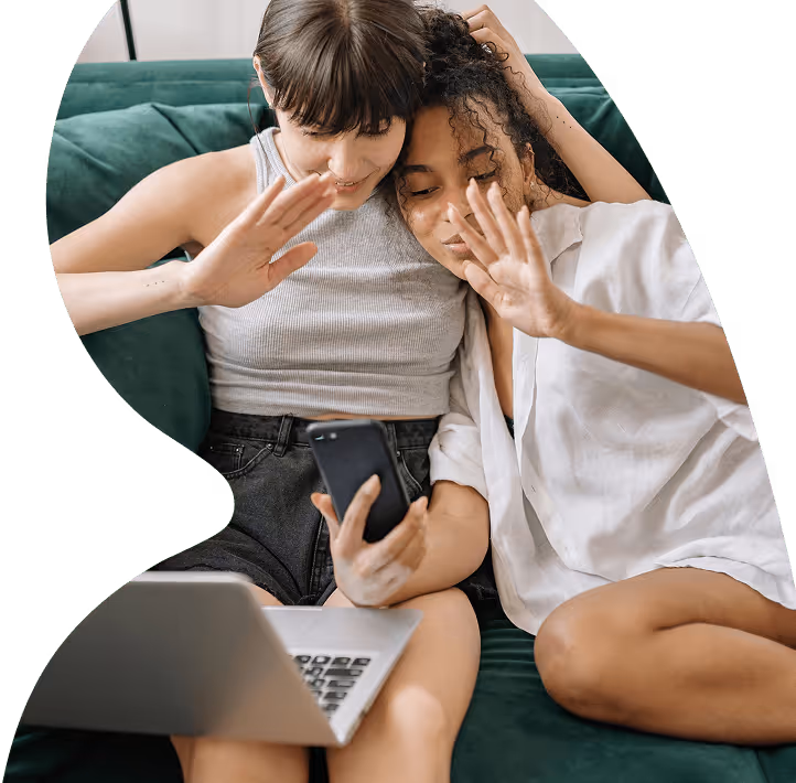 Two young women sitting on a couch waving at a smartphone during a video call with a laptop on their laps.