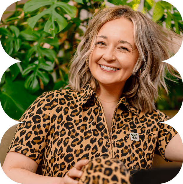 Smiling woman with shoulder-length blonde hair wearing a leopard print outfit sitting in front of green plants.