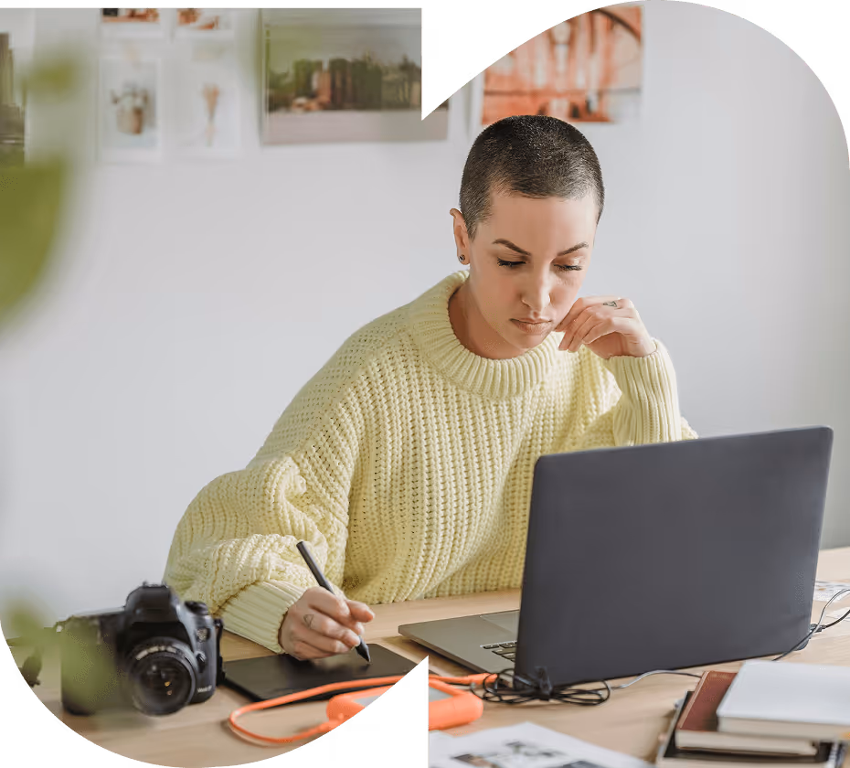 Person in a yellow sweater working on a laptop while using a digital drawing tablet at a wooden desk with a camera and books.