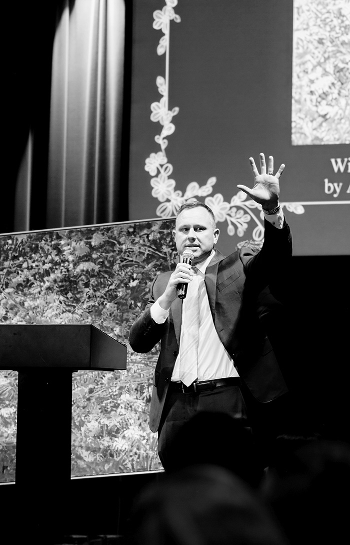 Man in suit speaking into a microphone with one hand raised on a stage during a presentation.