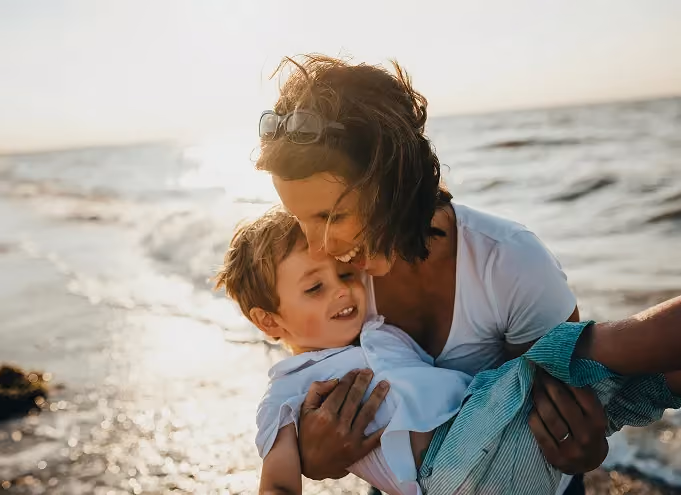 Smiling woman with sunglasses on her head holding and hugging a young boy near a sunlit ocean shore.