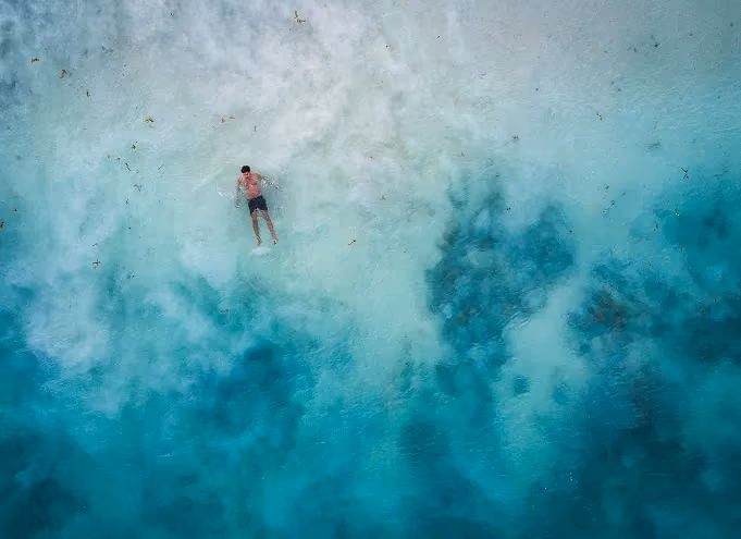 A man floating face down in clear, shallow turquoise water with visible underwater rocks and seaweed.