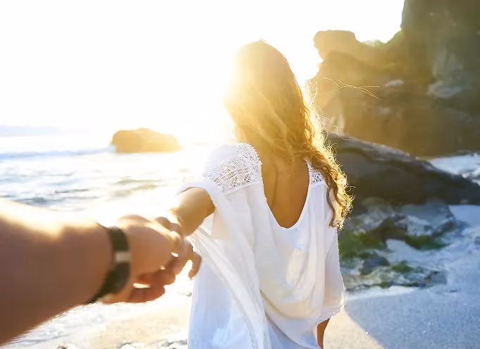 Woman in a white dress holding a man's hand leading him towards a sunlit seaside with rocky cliffs in the background.