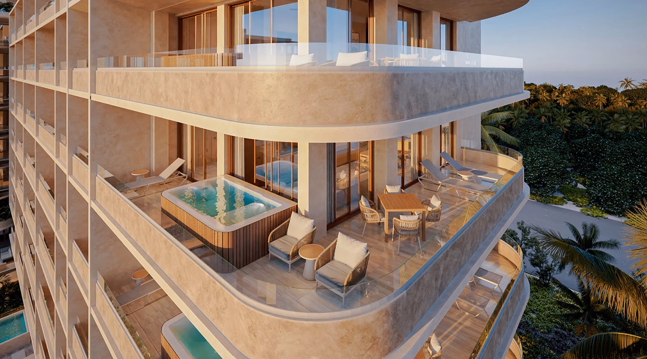 Corner balcony of a modern apartment with glass railing, featuring a hot tub, two lounge chairs, two cushioned armchairs with a small table, and a dining table with four chairs, overlooking lush greenery and palm trees during sunset.