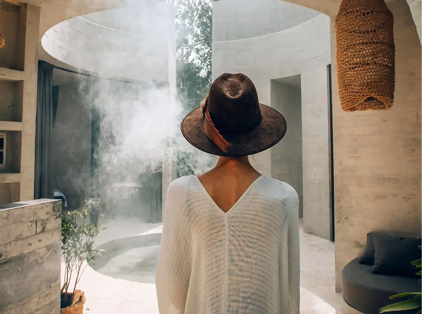 Person wearing a brown hat with a feather and a white textured sweater standing indoors with their back to the camera, facing a concrete circular open ceiling area with plants and a modern sofa.
