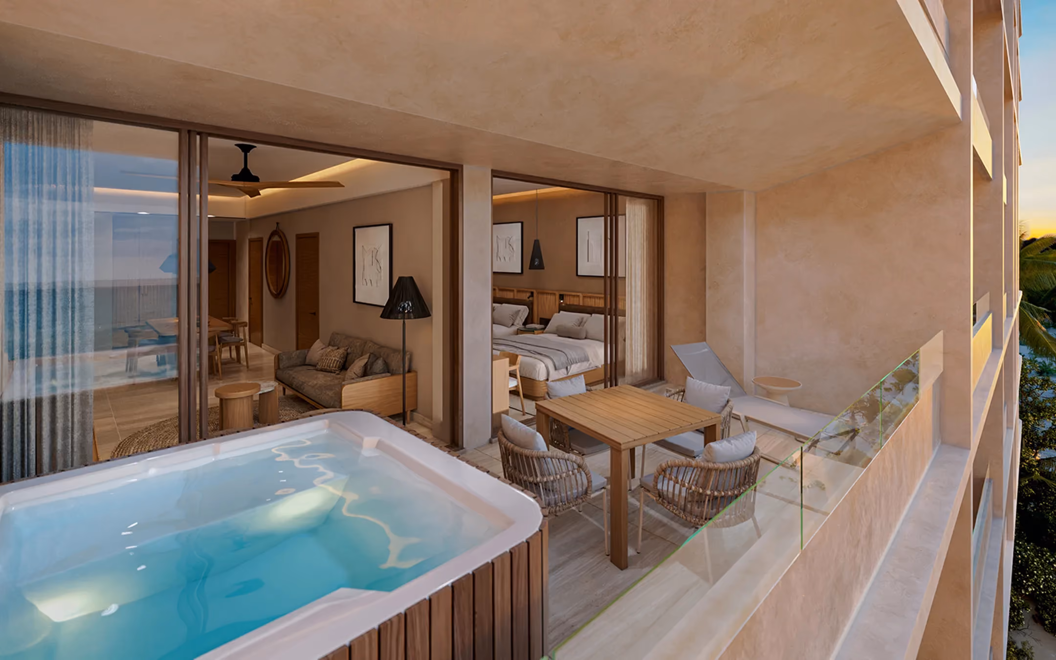 Balcony view of a modern hotel suite featuring a jacuzzi tub, wooden dining table with four cushioned wicker chairs, and a lounge chair, with glass railing overlooking palm trees and the ocean at sunset; inside, a living area with a sofa and a bedroom with a double bed are visible through large sliding glass doors.