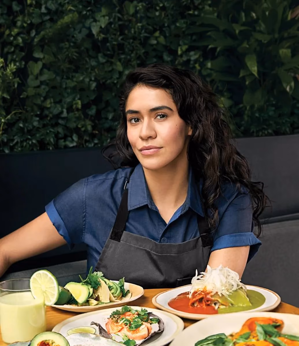 Woman with dark curly hair wearing a blue shirt and gray apron sitting at a table with a variety of colorful dishes including avocado slices, tacos, a glass with a lime slice, and plated meals with red and green sauces and fresh herbs.