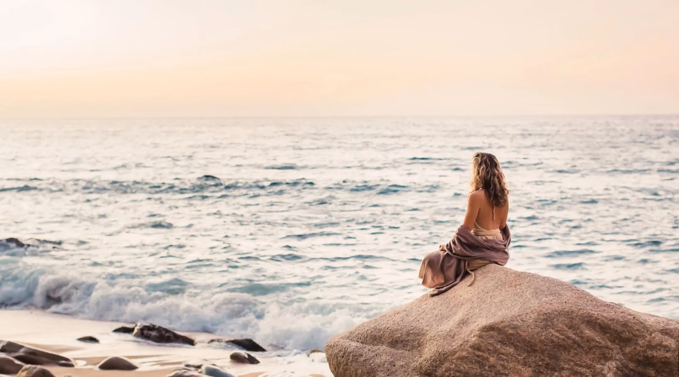 A woman with long curly hair, wrapped in a shawl, sits on a large rock facing the ocean at sunset, with gentle waves and a sandy shore with smaller rocks nearby.